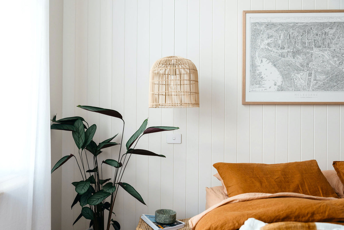 A large size open edition of The Melbourne Map black and white line drawing hanging in an oak frame on a white panelled wall. The picture is hanging above a bed that has burnt orange bed linen and is styled with rattan pendant lights and a lush, tall green palm