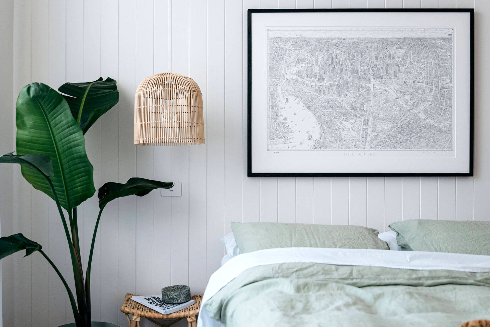 A limited edition of 1000 of The Melbourne Map line drawing. Framed in stained black Ash with a window mount. The print is hanging next to a rattan pendant light and above a bed with light green linen. There is a large palm to the left. 