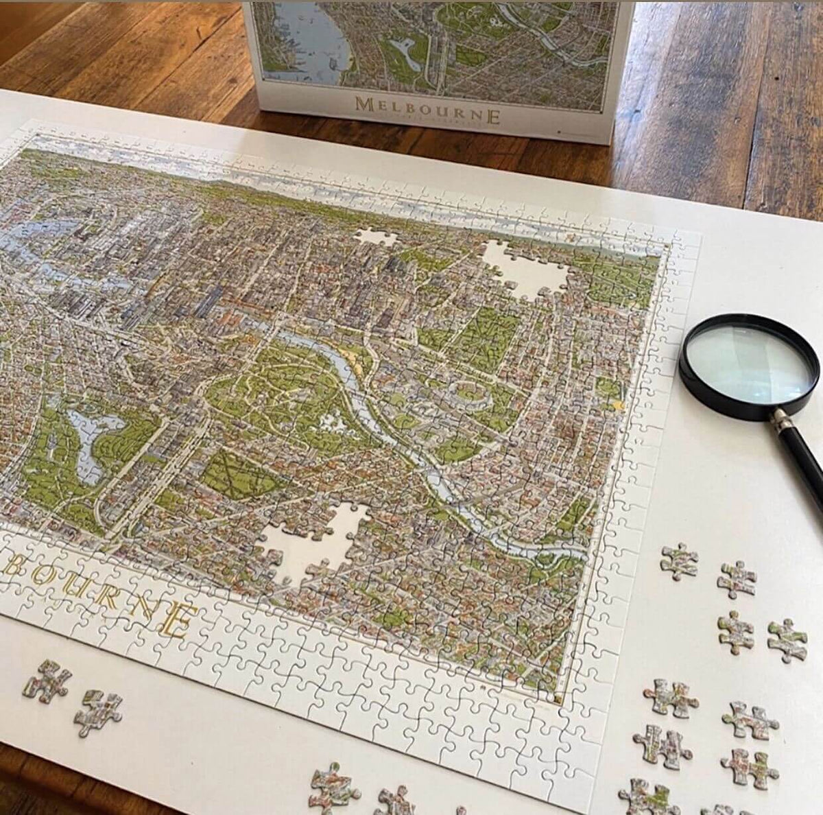 The Melbourne Map 1,000-piece jigsaw puzzle almost completed on a natural timber table with the box in the background. There are some loose pieces around the edge of the puzzle and a magnifying glass on the right. 