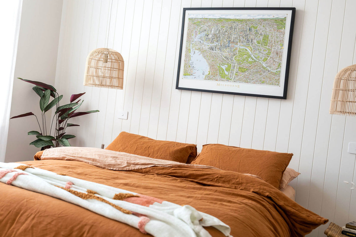 A large version of The Melbourne Map open edition art print in a black frame hanging above a bed with natural linen bedding and rattan pendant lights in an airy bright room