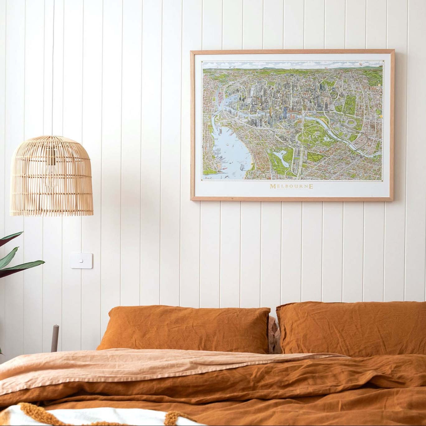 A large sized colour open edition of The Melbourne Map in an oak frame positioned above a bed with burnt orange linen and a rattan pendant light