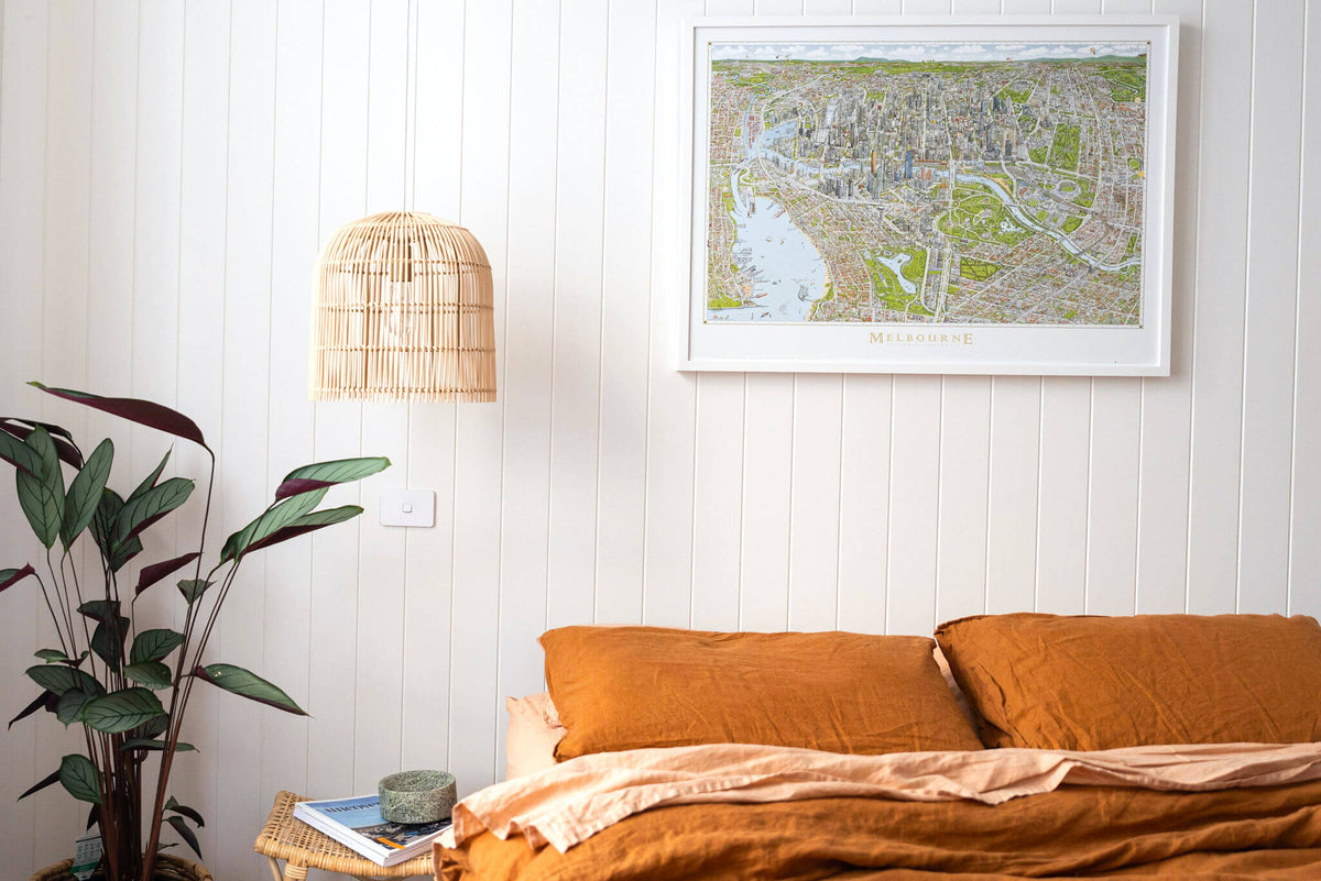 A copy of the large colour open edition of The Melbourne Map in a white frame positioned above a bed with burnt orange linen and a rattan pendant light