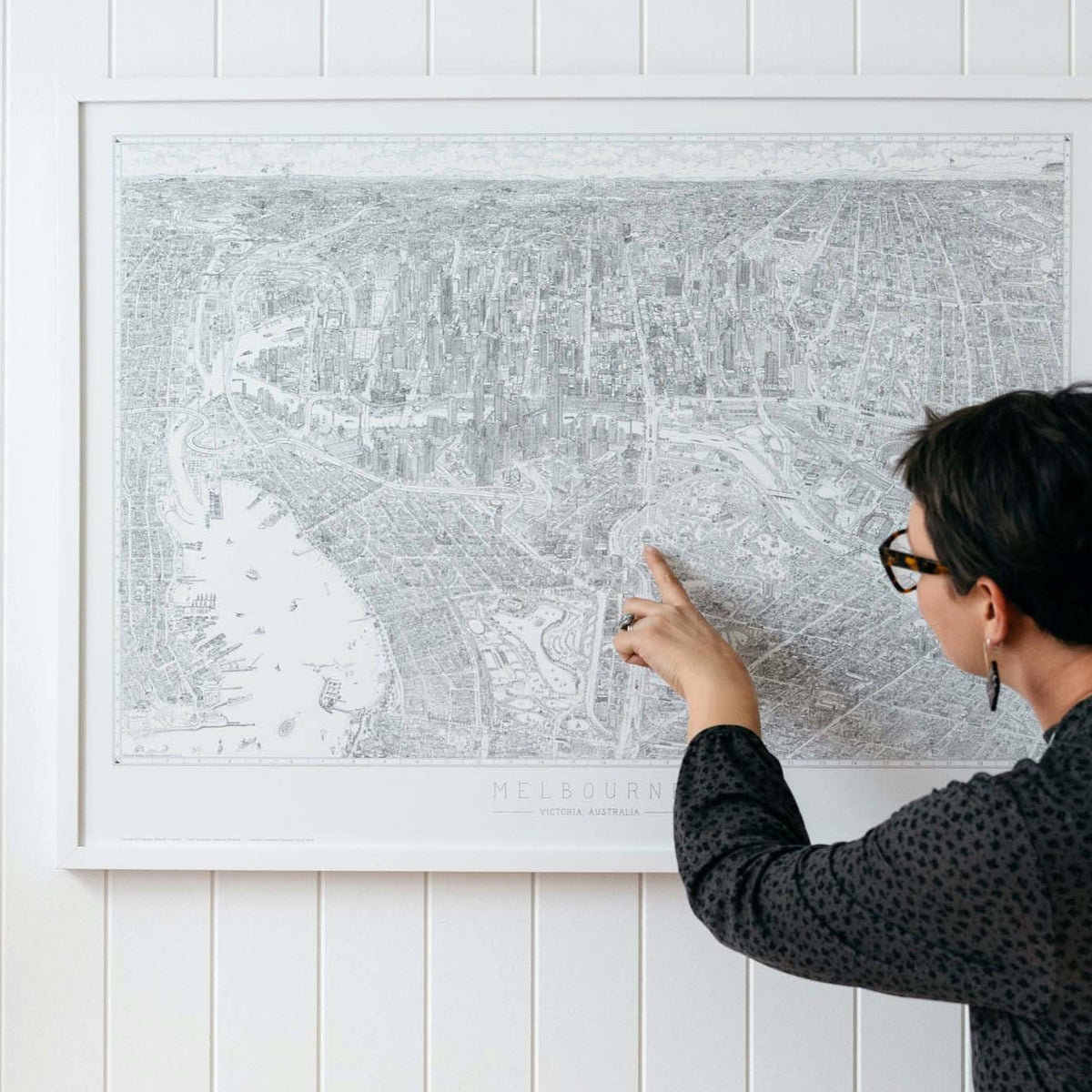 A woman wearing a dark green dress pointing at the Shrine of Remembrance on a large open edition of The Melbourne Map black and white line drawing which is in a white frame hanging in a bright airy room