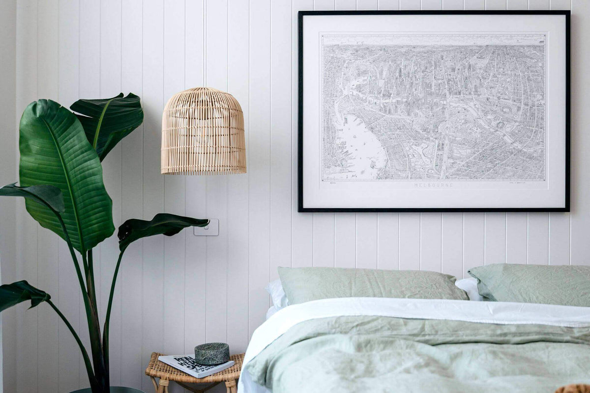 A black ash framed limited edition of 1000 version of The Melbourne Map black and white line drawing hanging above a bed with green linen on a white panelled wall with rattan pendant lights and a large palm plant 