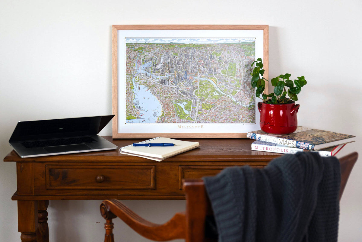 A small version of The Melbourne Map open edition colour art print in an oak frame sitting on a small timber console desk with laptop, books and plant. The antique chair has a knitted throw rug on it 