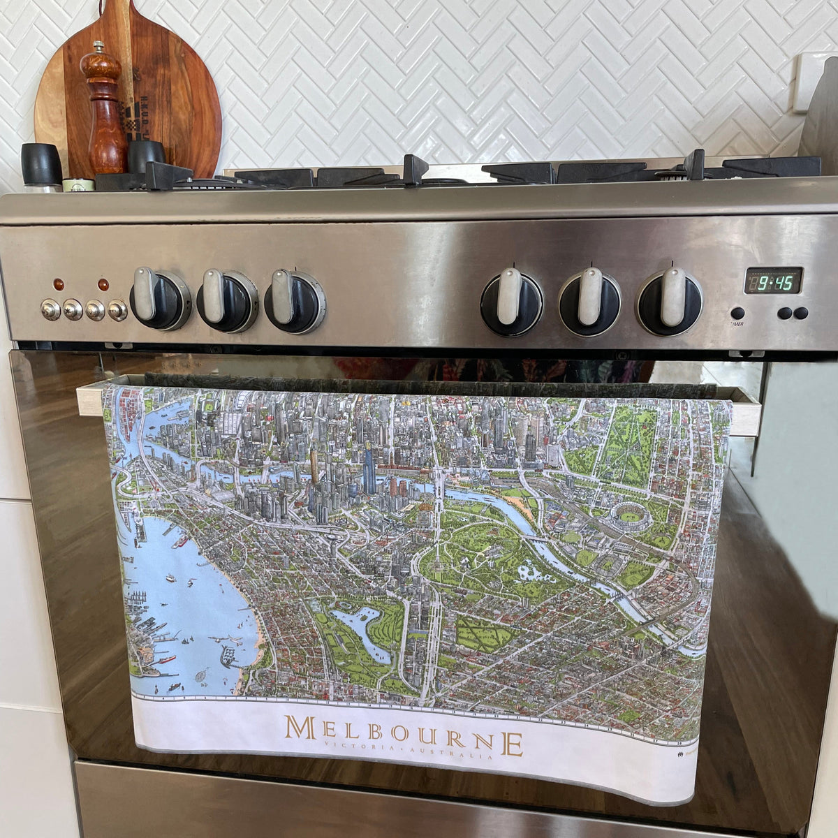 The Melbourne Map Tea Towel hanging on the handle of an oven door. The oven is a modern silver appliance with a white herringbone tiled backsplash. A wooden chopping board and pepper mill are in the background too. 