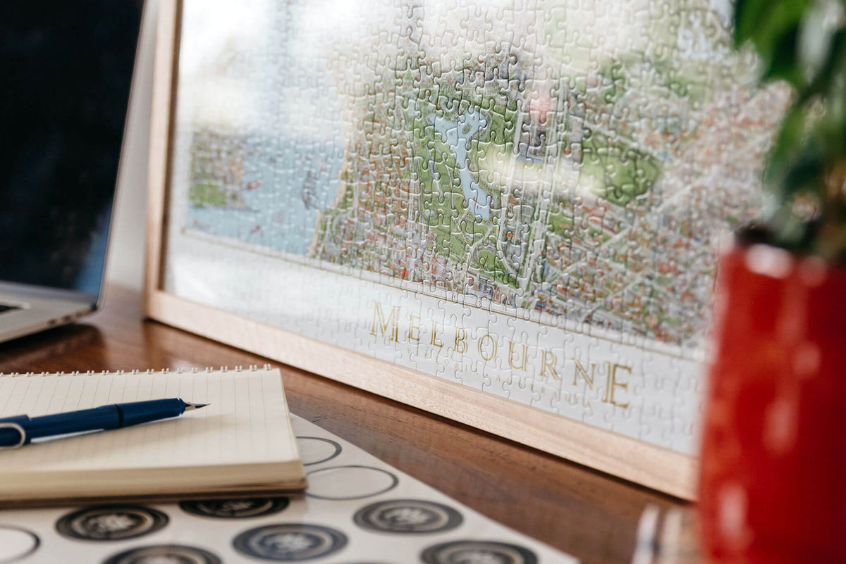 A framed, completed copy of The Melbourne Map 1,000-piece jigsaw puzzle in an oak frame sat on top of a natural timber desk with a red plant pot in the foreground and a notepad, fountain pen and some Melbourne Map logo stickers. 