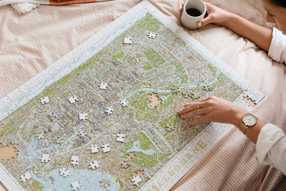 A young woman is laying on a bed which features orange pinstripe linen. She is working on The Melbourne Map 1,000-piece jigsaw puzzle while having a coffee. She is wearing a white linen shirt and has a gold watch on. 