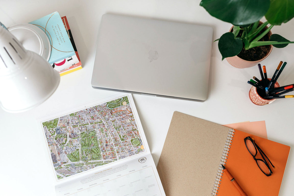 The &quot;Can You Find?&quot; calendar on a desk with an Apple laptop, pen pot, white lamp, orange notebook, small plant, and some books.