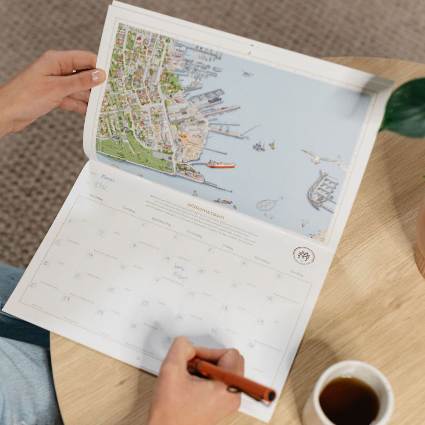 A Young woman is filling out the date sections on the Williamstown page of the "Can You Find?" perpetual calendar. She is sitting at a small oak table that has a plant on and a cup of coffee beside it.