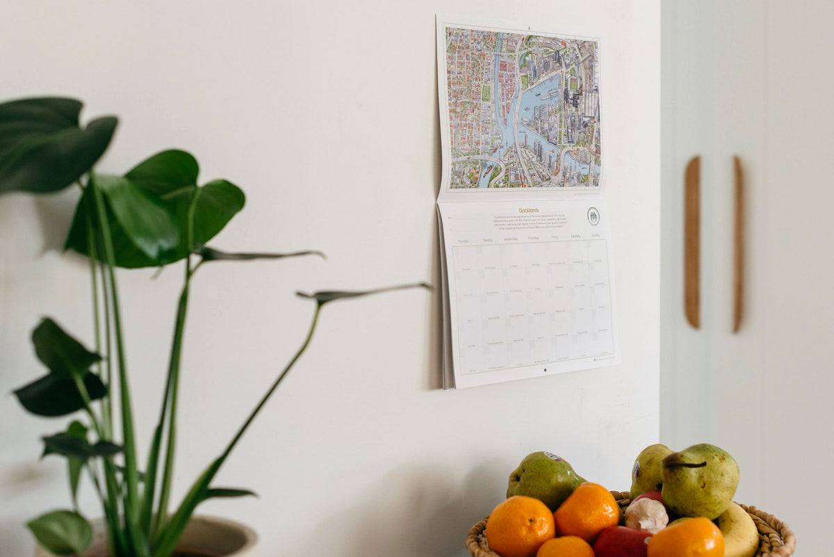 The &quot;Can You Find?&quot; calendar hanging on a kitchen wall. There is a rattan bowl of fruit below it and a small palm plant to the left of the image. 