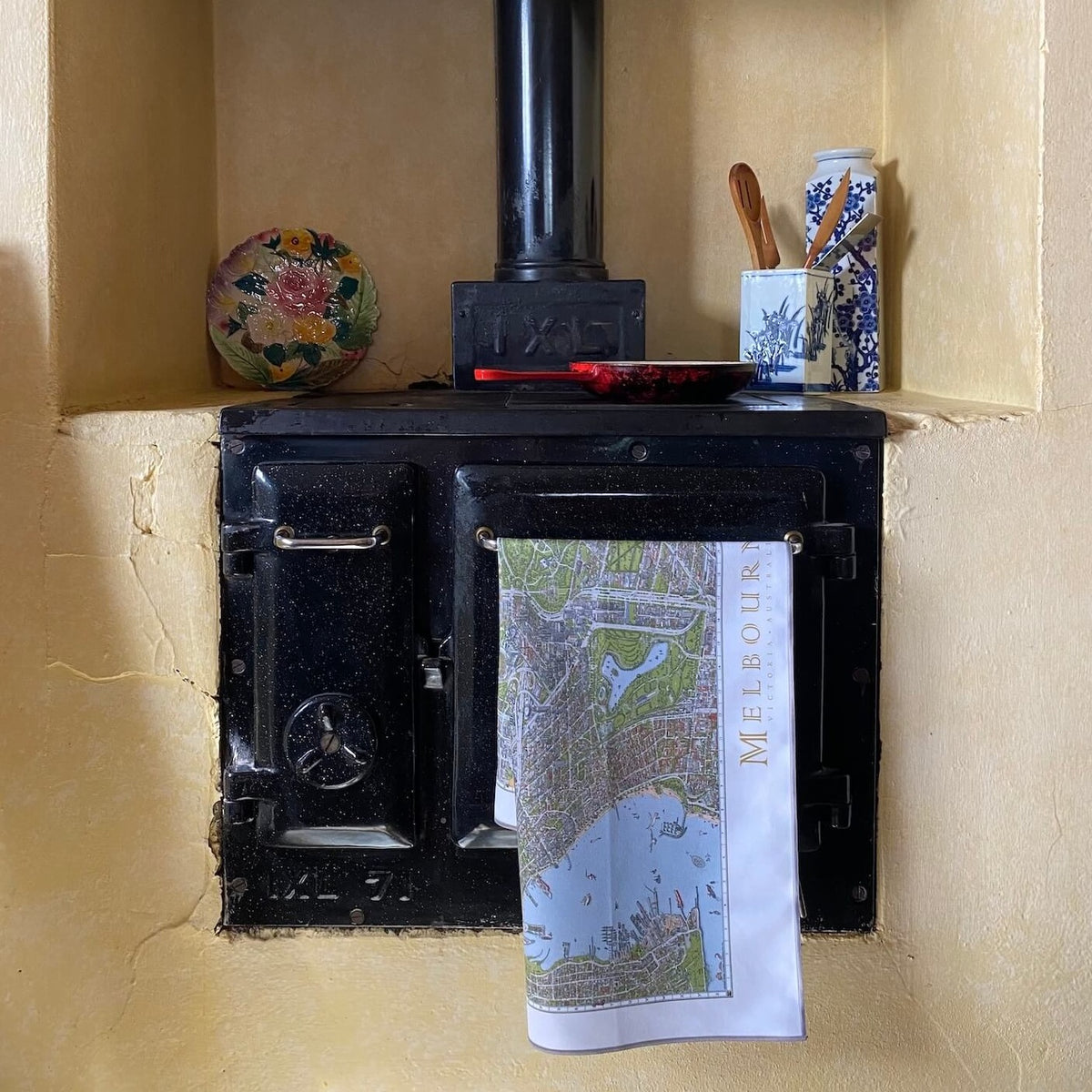 The Melbourne Map Tea Towel draping over the handle of an antique wood burning oven. There is a cast iron skillet on the stove with utensils pot and decorative plate either side. 