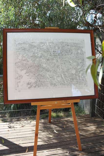 A copy of The 1990 Magic Map of Melbourne limited edition in a timber frame on an easel on display in an outdoor setting with trees in the background. 